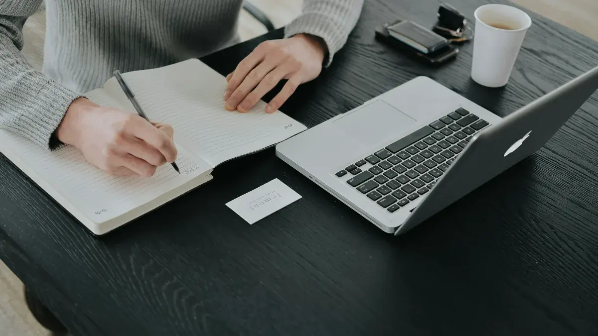 Self-employed professional writing notes beside a laptop at a home office desk